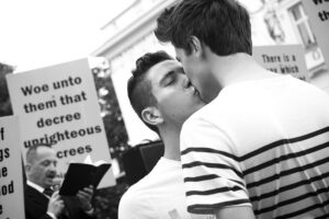 Two men kissing in front of a group of people holding signs.