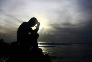A silhouette of a man sitting on a rock in front of the ocean.