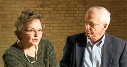 An older man and woman talking in front of a brick wall.