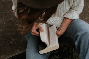 A woman wearing a hat and jeans reading a bible.