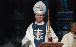 A priest in a white robe holding a book in front of a stained glass window.