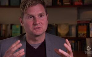 A man is sitting in front of a bookcase.