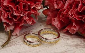 Two wedding rings on a table next to red flowers.