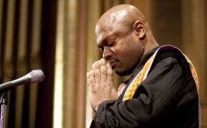 A black man praying in front of a microphone.