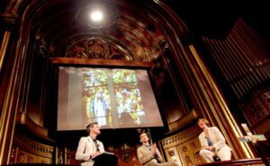 Three people sitting on a stage in front of a stained glass window.