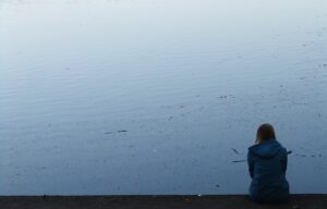 A woman sits on the edge of a lake looking at the water.