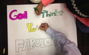 Two children writing on a sign that says god thinks you're fabulous.