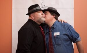 Two men in hats kissing in front of a sign.