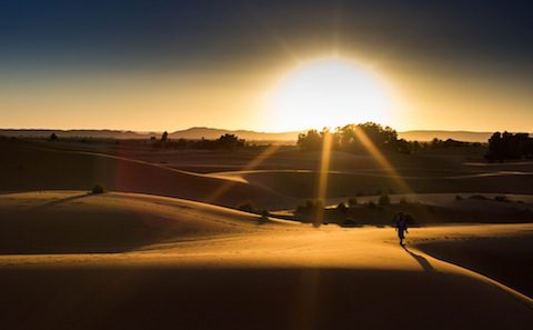 A man is walking through the sand dunes at sunset.