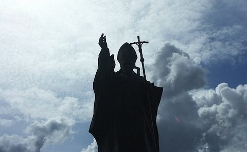 A statue of a man with a cross in front of a cloudy sky.