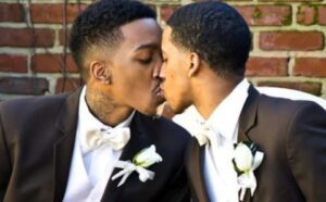 Two men in suits kissing in front of a brick wall.