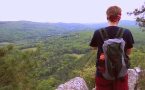 A man with a backpack is standing on top of a cliff overlooking a valley.