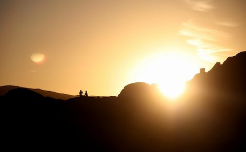 A silhouette of two people standing on top of a rock at sunset.