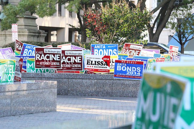 A group of campaign signs on a sidewalk in front of a building.