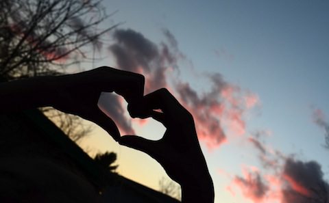 A person's hand making a heart shape at sunset.