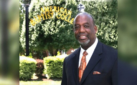 A man in a suit and orange tie is standing in front of a sign.