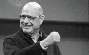 A black and white photo of a bald man giving a thumbs up.