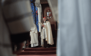 A priest is standing in front of a stained glass window.