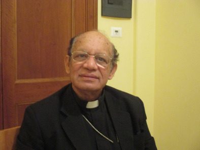 A priest in a black suit sitting in front of a wooden table.