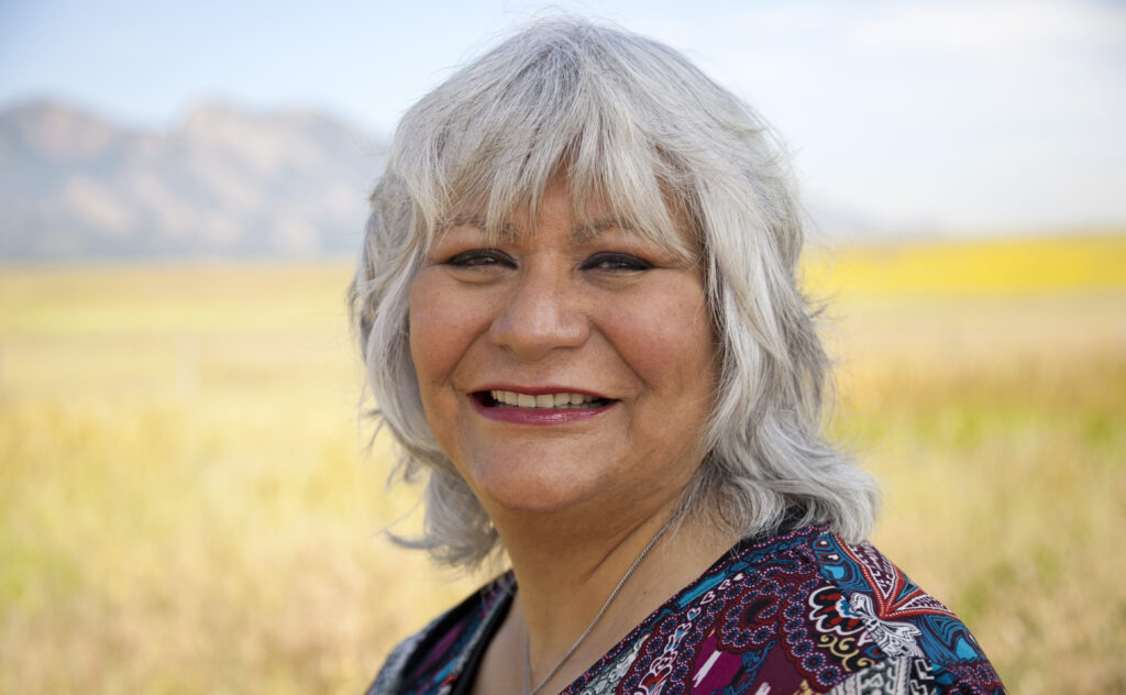 An older woman smiling in a field with mountains in the background.