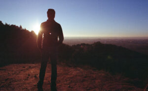 A man standing on top of a hill with the sun setting behind him.