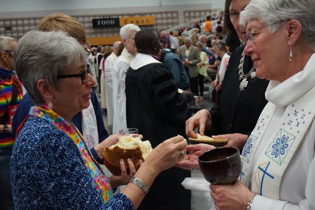 Two women in white robes are holding bread in front of a crowd.