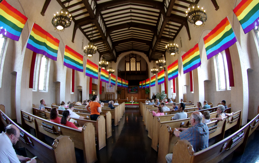 People sitting in a church with rainbow flags hanging from the ceiling.