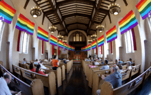 People sitting in a church with rainbow flags hanging from the ceiling.