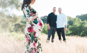 A pregnant woman in a floral dress is standing in a field with her husband.