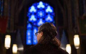 A woman in glasses looking at a blue stained glass window.