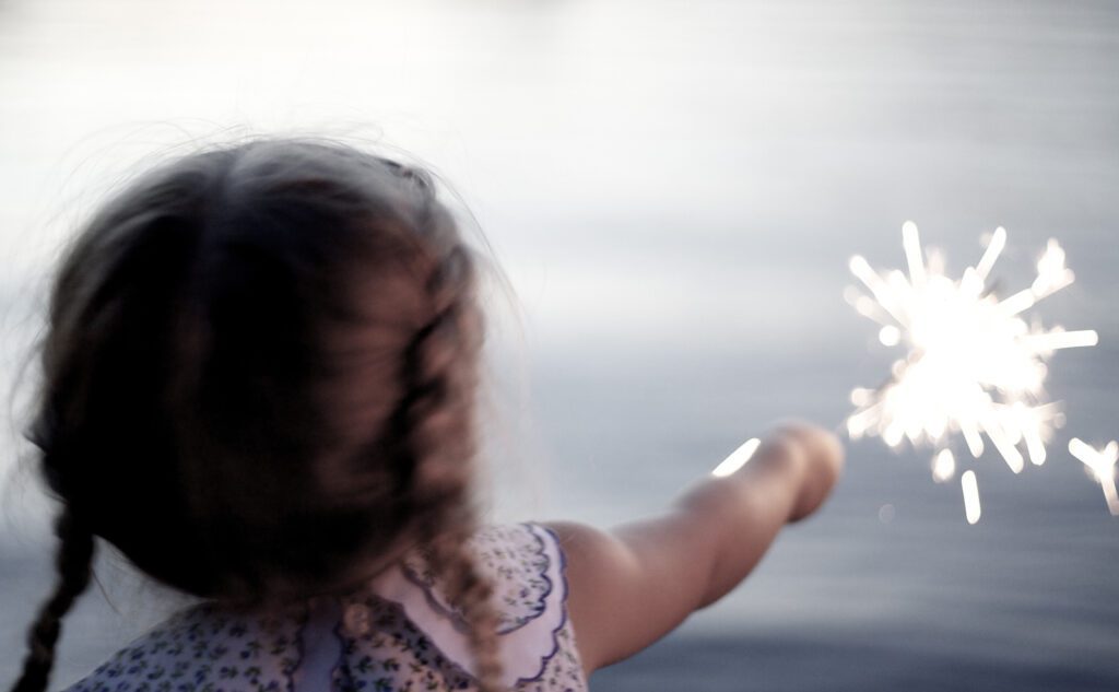 A little girl holding a sparkler in front of a body of water.