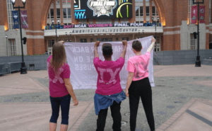 Three women in pink shirts holding a banner in front of a building.