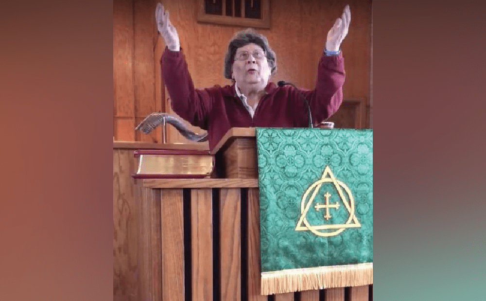 A woman standing at a pulpit with her hands raised.