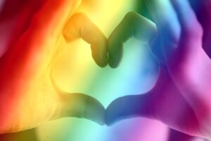 Two hands making a heart shape with a rainbow background.
